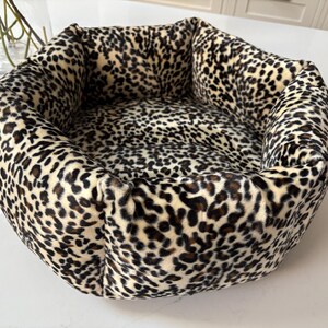 May include: A plush pet bed with a leopard print pattern. The bed is round with a cushioned rim and a soft interior. The colors are cream, black, and brown. The bed is designed for small to medium-sized pets.