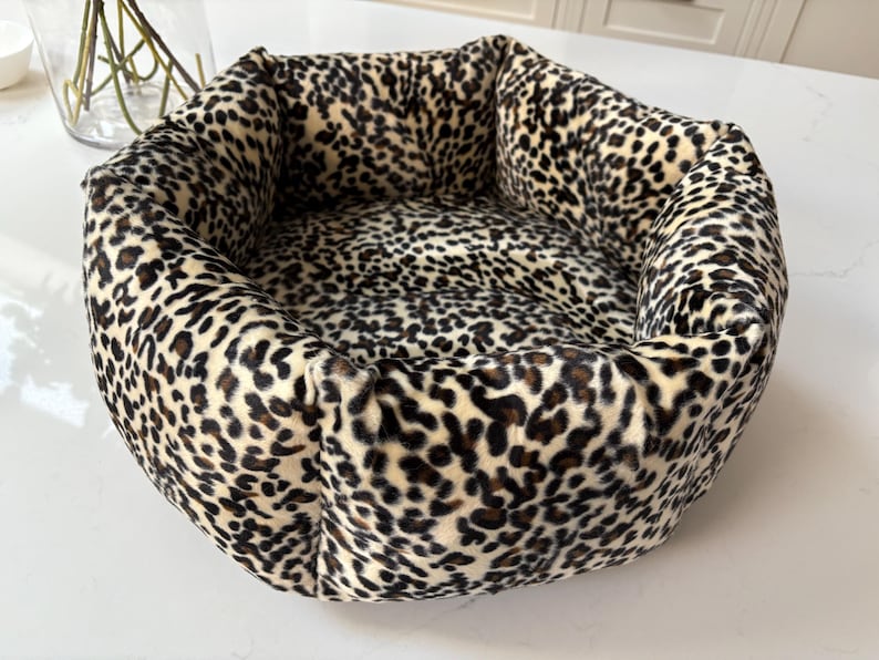 May include: A plush pet bed with a leopard print pattern. The bed is round with a cushioned rim and a soft interior. The colors are cream, black, and brown. The bed is designed for small to medium-sized pets.