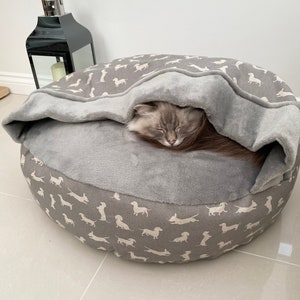 May include: A grey and white dog-print pet bed with a grey fleece liner. A cat is sleeping inside the bed under the liner.