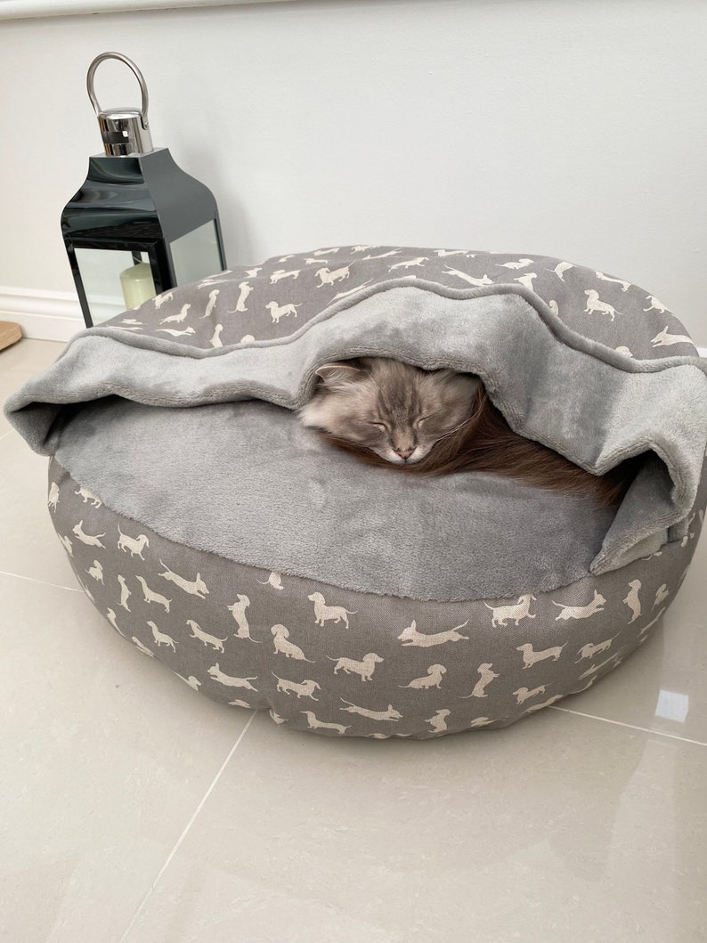 May include: A grey and white dog-print pet bed with a grey fleece liner. A cat is sleeping inside the bed under the liner.