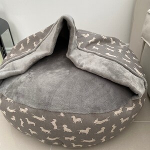 May include: A gray and white dog bed with a removable cover. The cover has a pattern of white dachshunds on a gray background. The bed has a soft, plush lining.