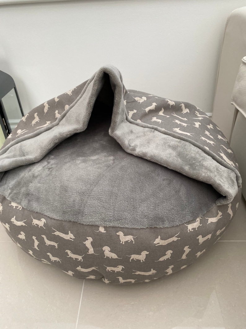 May include: A gray and white dog bed with a removable cover. The cover has a pattern of white dachshunds on a gray background. The bed has a soft, plush lining.