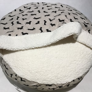 May include: A round pet bed with a removable, plush, white sherpa liner. The outer fabric is beige with a black dachshund print.