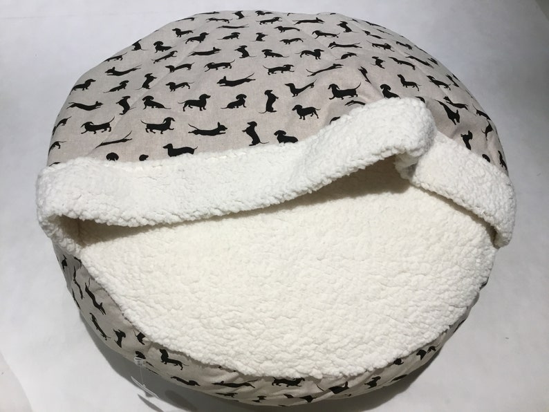 May include: A round pet bed with a removable, plush, white sherpa liner. The outer fabric is beige with a black dachshund print.