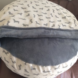 May include: A round pet bed with a grey fleece lining and a beige outer fabric featuring a repeating pattern of dachshund silhouettes.