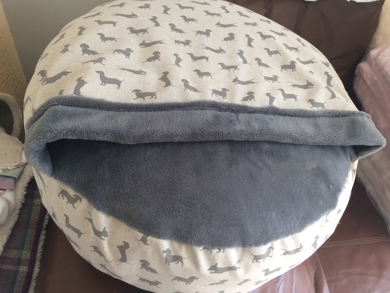May include: A round pet bed with a grey fleece lining and a beige outer fabric featuring a repeating pattern of dachshund silhouettes.
