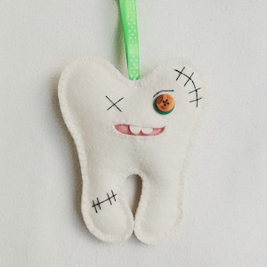 May include: White felt tooth with a stitched-on button eye, a stitched-on mouth, and black stitching details. The tooth is hanging from a green ribbon with white polka dots.