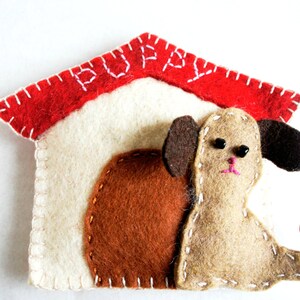 May include: A felt dog house with a red roof and the word "Puppy" stitched on the roof. The dog house is white and has a brown felt dog inside.