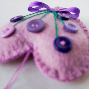 May include: A pink felt heart-shaped pincushion with purple buttons and green thread. The pincushion has a purple ribbon bow on top.