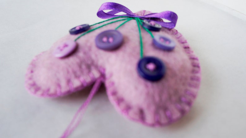 May include: A pink felt heart-shaped pincushion with purple buttons and green thread. The pincushion has a purple ribbon bow on top.