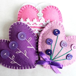 May include: Three felt heart-shaped ornaments with decorative stitching. Two are purple with purple buttons and embroidered flowers. One is pink with white lace and a pink button.