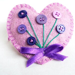 May include: A pink felt heart with a purple ribbon and five buttons sewn on to look like flowers. The buttons are purple, blue, and light pink.