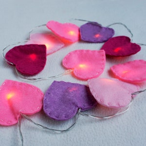 May include: A string of fairy lights with felt heart-shaped covers in pink, purple, and red. The lights are lit and glowing.