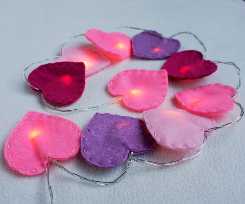 May include: A string of fairy lights with felt heart-shaped covers in pink, purple, and red. The lights are lit and glowing.