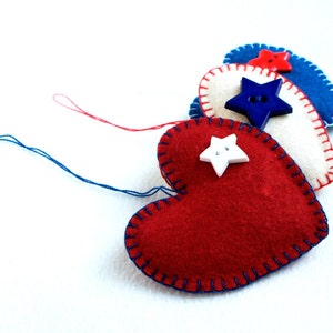 May include: Handmade felt heart ornaments in red, white, and blue, with star-shaped button embellishments. The hearts are stitched with contrasting thread and have hanging threads. These patriotic decorations are perfect for Independence Day.