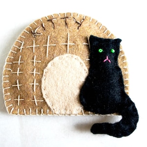 May include: A felt cat with green eyes and a pink nose sitting in a brown felt house with the word "Kitty" stitched on the top.