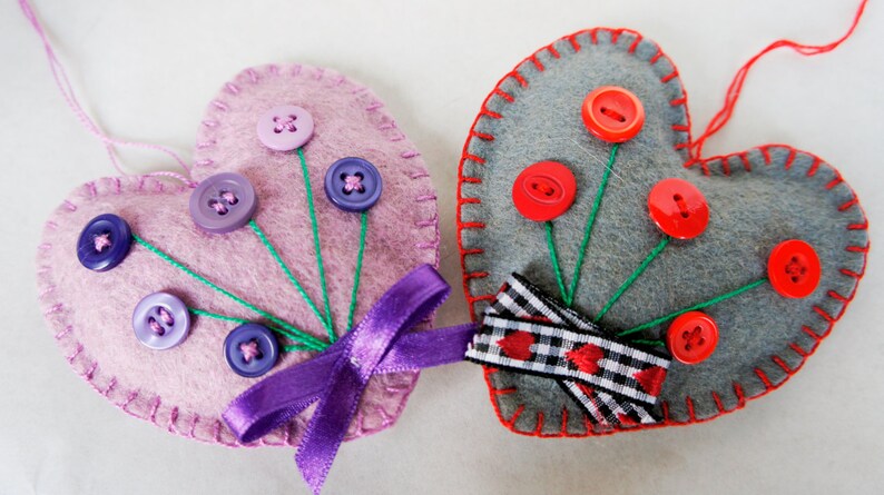 May include: Two felt heart ornaments with button embellishments. One heart is pink with purple buttons and a purple ribbon. The other heart is gray with red buttons and a black and white checkered ribbon.