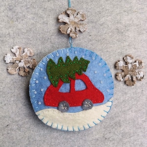 May include: A felt Christmas ornament depicting a red car carrying a green Christmas tree on a blue background with white snowflakes.