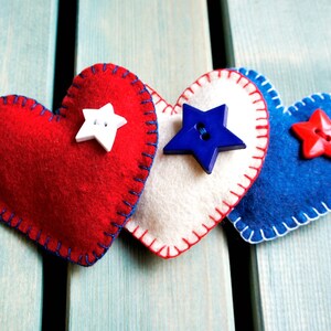 May include: Three felt heart decorations in red, white, and blue, with star-shaped button embellishments. Each heart is hand-stitched with contrasting thread. The hearts are arranged on a light blue wooden surface.