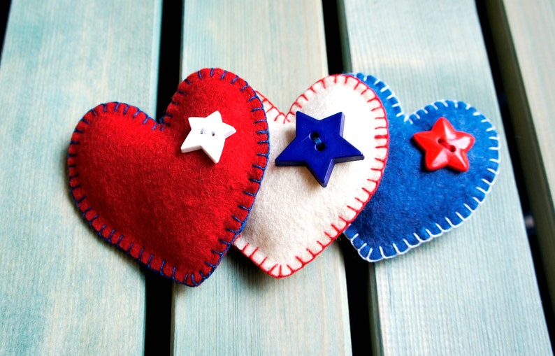 May include: Three felt heart decorations in red, white, and blue, with star-shaped button embellishments. Each heart is hand-stitched with contrasting thread. The hearts are arranged on a light blue wooden surface.
