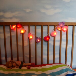 May include: A wooden crib with a white and blue striped mattress and a string of heart-shaped lights hanging above it. The lights are pink and red and are lit up.