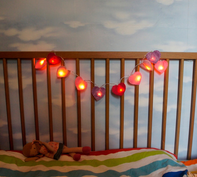 May include: A wooden crib with a white and blue striped mattress and a string of heart-shaped lights hanging above it. The lights are pink and red and are lit up.