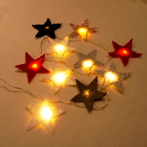 Può includere: Una catena di luci fatate con coperture in tessuto a forma di stella. Le luci sono bianche e le stelle sono di vari colori, tra cui rosso, bianco e nero.