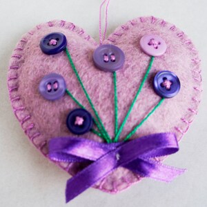 May include: A pink felt heart with a purple satin ribbon and purple, pink, and blue buttons sewn on to look like flowers.