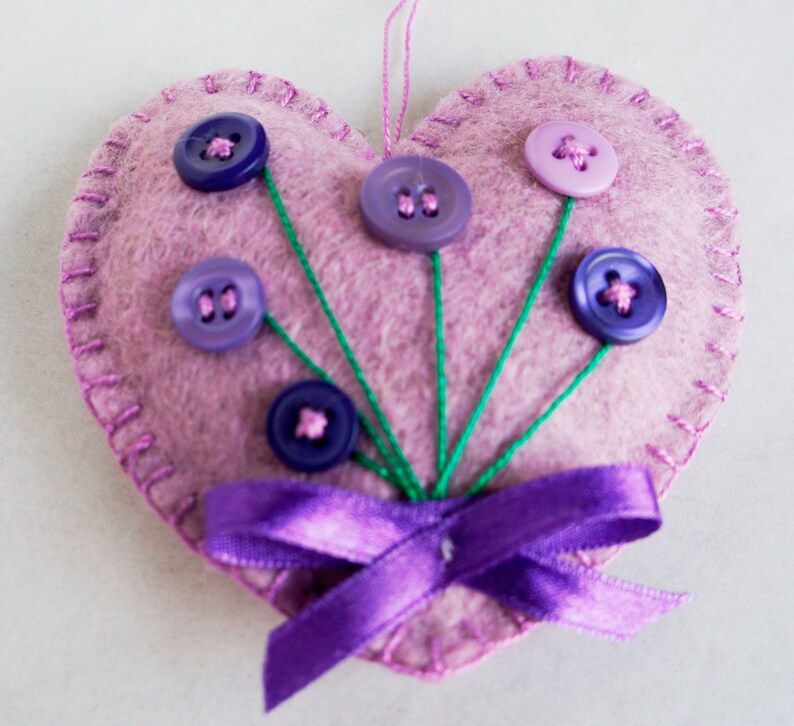 May include: A pink felt heart with a purple satin ribbon and purple, pink, and blue buttons sewn on to look like flowers.