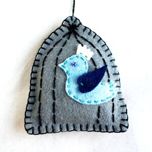 May include: A gray felt birdhouse ornament with a blue bird with a white crown and a blue wing inside. The birdhouse has black stitching around the edges.