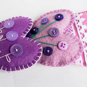 May include: Three felt heart-shaped pincushions with decorative stitching and buttons. The pincushions are purple, pink, and lavender.