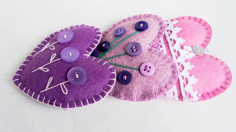 May include: Three felt heart-shaped pincushions with decorative stitching and buttons. The pincushions are purple, pink, and lavender.