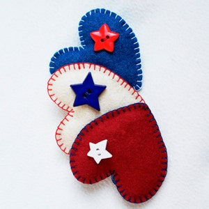 May include: Three felt heart decorations in red, white, and blue, stitched with contrasting thread. Each heart features a star-shaped button in a coordinating color. The hearts are arranged in a row, evoking a patriotic theme.