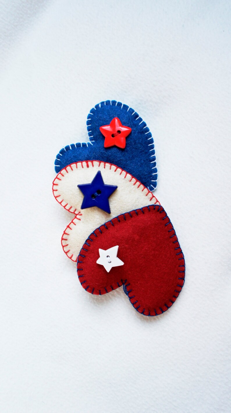 May include: Three felt heart decorations in red, white, and blue, stitched with contrasting thread. Each heart features a star-shaped button in a coordinating color. The hearts are arranged in a row, evoking a patriotic theme.