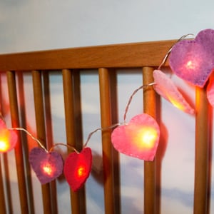 May include: A string of fairy lights with heart-shaped felt covers in shades of pink and purple. The lights are strung across a wooden crib rail.