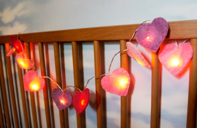 May include: A string of fairy lights with heart-shaped felt covers in shades of pink and purple. The lights are strung across a wooden crib rail.