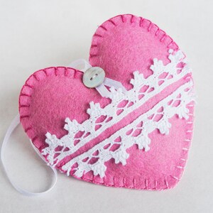 May include: A pink felt heart with white lace trim and a pearl button. The heart has a white ribbon loop for hanging.
