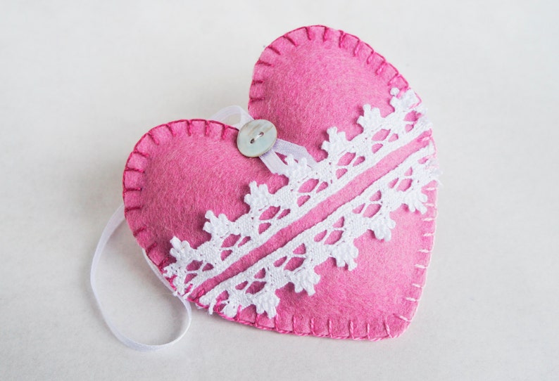 May include: A pink felt heart with white lace trim and a pearl button. The heart has a white ribbon loop for hanging.