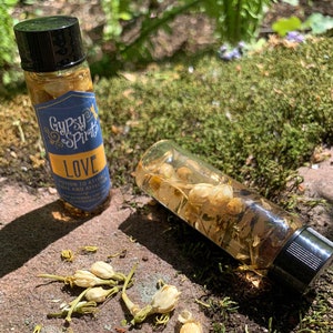 May include: Two small glass bottles with cork stoppers and labels that say "Gypsy Spirits Love Potion to Attract Love and Affection". The bottles are filled with a clear liquid and white flower petals. The bottles are sitting on a bed of moss.