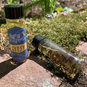May include: Two small glass bottles with black caps. The bottle on the left is labeled "Gypsy Spirits" and "Gypsy Potion for Strength, Spirit and Success". The bottle on the right is filled with a clear liquid with gold flakes.
