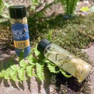 May include: Two small glass bottles with black caps. One bottle has a blue and yellow label that reads "Gypsy Spirits Luck". The other bottle is filled with clear liquid and white flowers. The bottles are surrounded by green ferns and moss.