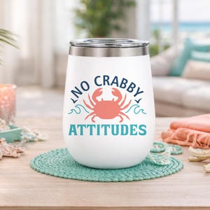 Puede incluir: Vaso de vino aislado blanco con tapa plateada, con el texto "NO CRABBY ATTITUDES" y un dise&ntilde;o de cangrejo coral. El vaso se encuentra sobre un posavasos turquesa, con decoraci&oacute;n de tem&aacute;tica playera en el fondo.