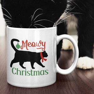 May include: White ceramic mug with a black cat silhouette holding a green Christmas ornament. The text "Meowy Christmas" is printed on the mug in red and green lettering.