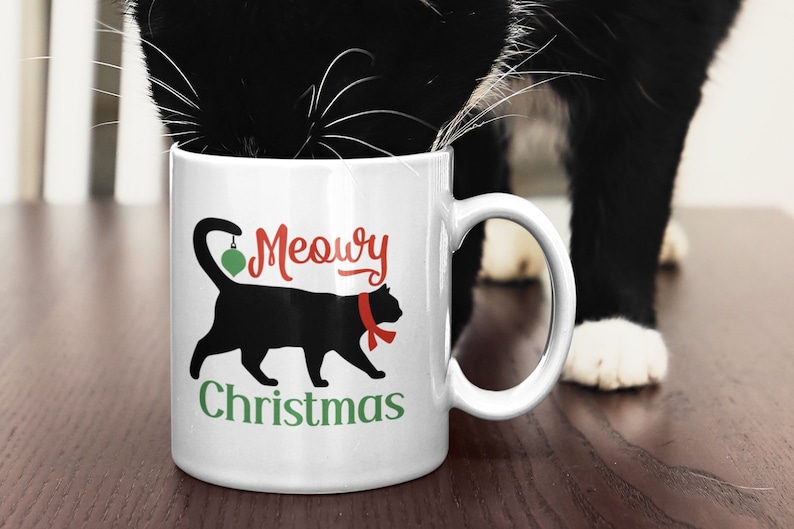 May include: White ceramic mug with a black cat silhouette holding a green Christmas ornament. The text "Meowy Christmas" is printed on the mug in red and green lettering.