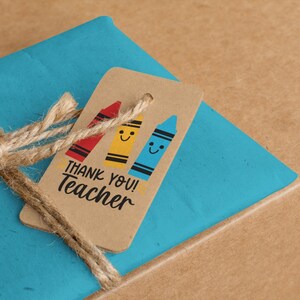 May include: A gift wrapped in blue paper, tied with twine, and a brown tag. The tag features three smiling crayon illustrations in red, yellow, and blue, with the text "THANK YOU! Teacher".