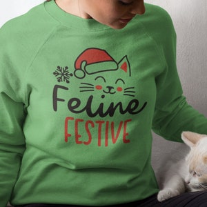 May include: A green sweatshirt with a festive cat wearing a Santa hat and the text "Feline Festive".