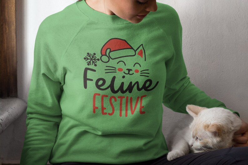 May include: A green sweatshirt with a festive cat wearing a Santa hat and the text "Feline Festive".
