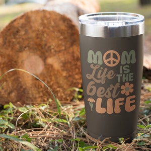 May include: A black stainless steel tumbler with a peace sign and the text "Mom Life is the Best Life" in a retro font. The tumbler is sitting on a brown log in a grassy area.