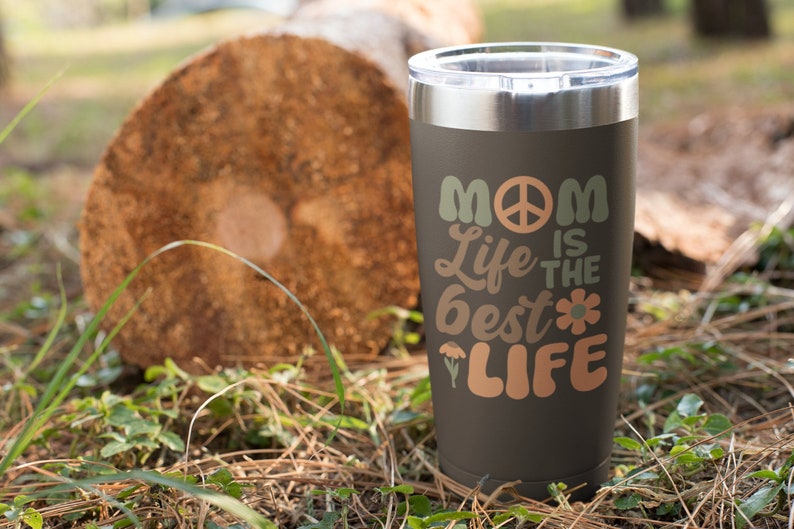 May include: A black stainless steel tumbler with a peace sign and the text "Mom Life is the Best Life" in a retro font. The tumbler is sitting on a brown log in a grassy area.