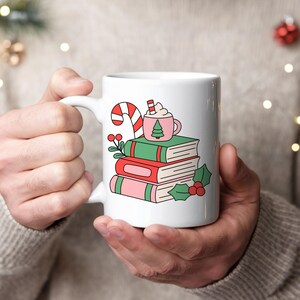 May include: A white ceramic mug featuring a colourful graphic of a stack of three books in green, red, and pink. A small pink mug with a green tree icon, a candy cane, and holly berries with green leaves are illustrated on top of the books.